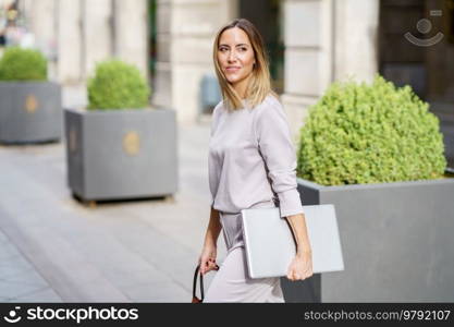 Side view of self assured adult female entrepreneur with blond hair in classy clothes, smiling and looking away while walking on city street with laptop in hand. Smiling businesswoman with laptop crossing road in city modern district