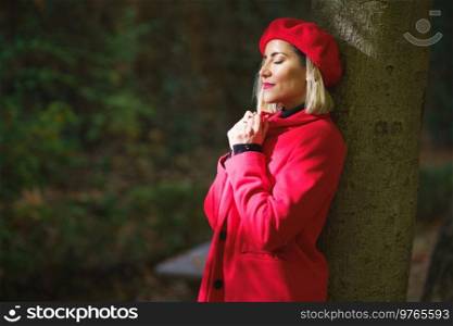 Side view of serene female with closed eyes adjusting collar of red coat while standing in sunlight near tree in park. Gentle woman in red beret and coat leaning on tree