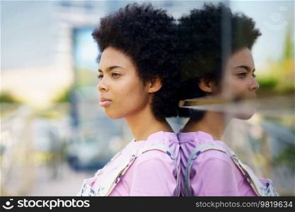 Side view of serious African American female with Afro hairstyle standing near glass house on street against blurred background in city. Black woman near glass building in city