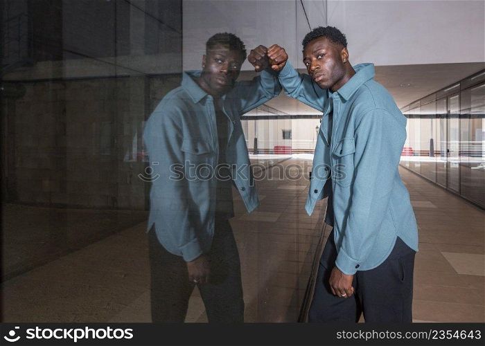 Side view of serious young black male model in trendy outfit leaning on glass mirrored wall and looking at camera while standing in hallway of modern underground. Stylish African American guy leaning on reflecting glass wall