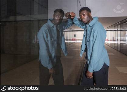 Side view of serious young black male model in trendy outfit leaning on glass mirrored wall and looking at camera while standing in hallway of modern underground. Stylish African American guy leaning on reflecting glass wall