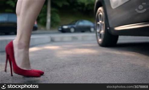 Side view of sexy female legs in red high heels getting into car. Beautiful woman with perfect slim legs wearing red classic high heel shoes opening car door and getting into car in daylight. Low section.