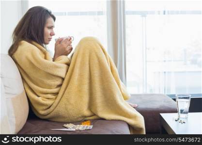 Side view of sick woman having coffee on sofa in living room