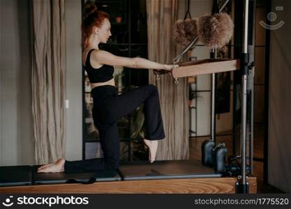 Side view of slender redhead female pilates trainer in black tight sportswear showing legs stretching exercises during training session in fitness studio, fit woman exercising in Cadillac reformer. Slender fit pilates trainer showing stretching exercises on cadillac reformer