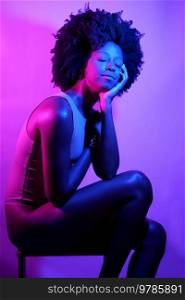 Side view of slim African American female model in bodysuit, with curly hair touching shoulder and looking away while sitting on stool under violet neon light. Positive black woman under neon light with eyes closed.