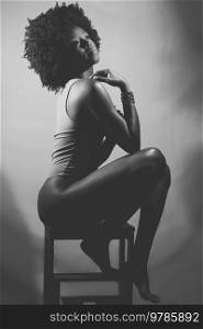 Side view of slim African American female model in bodysuit, with curly hair touching shoulder and looking away while sitting on stool. Black and white photograph.. Black and white photograph of sensual black woman under neon light