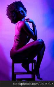 Side view of slim African American female model in bodysuit, with curly hair touching shoulder and looking away while sitting on stool under violet neon light. Sensual black woman under neon light