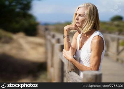 Side view of slim aged female standing near railing on wooden boardwalk on seashore in summer and looking away in contemplation. Thoughtful senior woman on boardwalk on beach