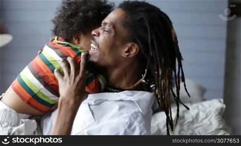 Side view of smiling african dad lying on his back on the bed and having fun with his adorable mixed race toddler son. Playful handsome father with dreadlocks playing with his boy, lifting him up in the air and relaxing together at home.