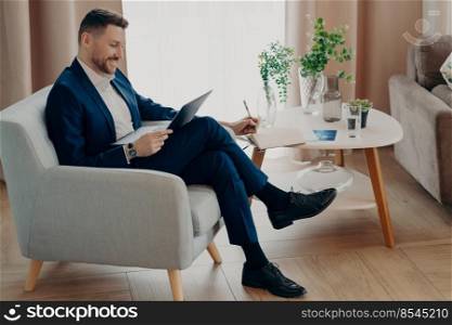 Side view of smiling bearded businessman in suit watching online business video while sitting in armchair and working remotely, using laptop and studying online. Business people lifestyle concept. Happy young businessman enjoying working at home in living room