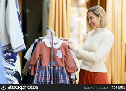 Side view of smiling female customer choosing apparel for girl while standing near rack with various stylish clothes in modern boutique. Content woman choosing clothes for little girl