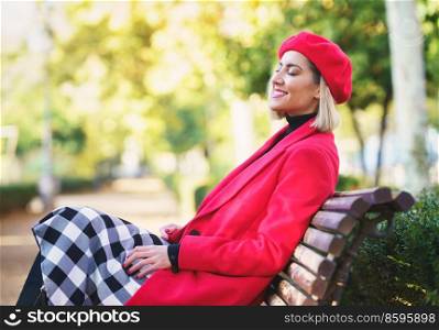 Side view of smiling female in red coat and beret sitting on wooden bench with closed eyes in autumn park on blurred background. Positive woman sitting on bench in park