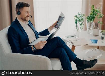 Side view of smiling manager using headset for video call with colleagues and looking at laptop screen while sitting in an armchair in the living room of his apartment, discussing project online. Businessman in suit discussing report with team online
