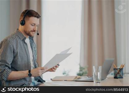 Side view of smiling young man freelancer looking at documents and feeling satisfied while sitting at table and using laptop. Guy holding papers and working remotely at home. Bearded male freelancer holding documents and working remotely at home