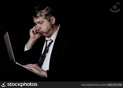 Side view of stressed business man on cell phone while looking at computer screen. Dark background with light on subject.