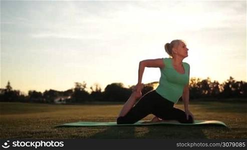 Side view of stunning adult sporty woman practicing traditional yoga pose in the park, doing pigeon yoga pose on green law in glow of sunset. Fit sporty lady in sportswear stretching in one legged pigeon pose while working out outdoors. Dolly shot.