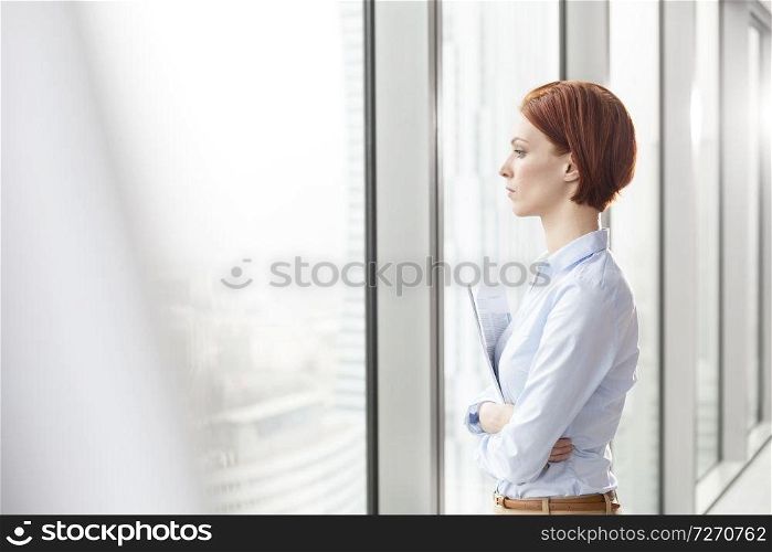 Side view of thoughtful young businesswoman holding documents while looking through window at office