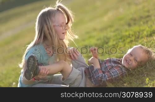 Side view of two playful children having fun on green field in park. Lovely blonde sister in eyeglasses kissing belly of her toddler brother, kids fooling around and laughing. Excited siblings enjoying time togehter outdoors.