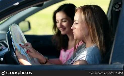 Side view of two pretty girls sitting in the car and paving the travel route on the map with finger. Young beautiful women on vacation are discussing directions of their roadtrip.