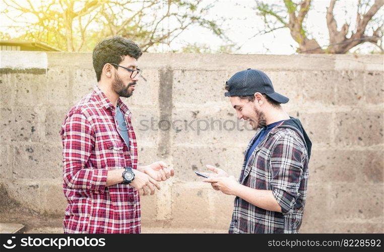 Side view of two young friends chatting outdoors, two friends chatting while smiling and using cellphone. View of two teenagers talking face to face while using the cell phone