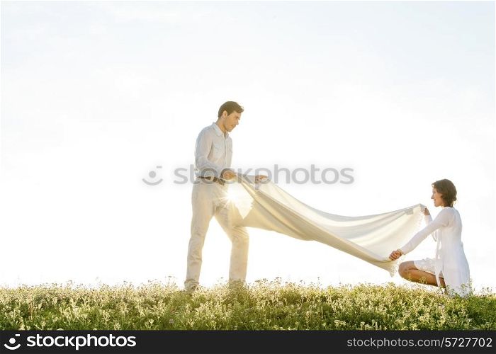 Side view of woman and man spreading picnic blanket on grass during sunny day