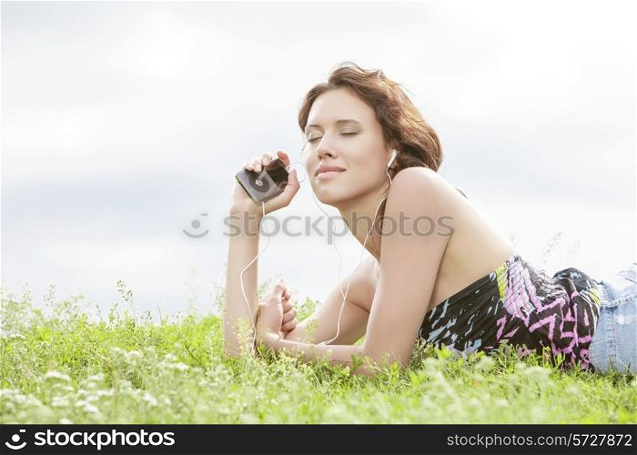 Side view of woman listening to music through MP3 player using headphones while lying on grass against sky