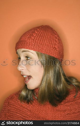 Side view of young adult Caucasian woman on orange background wearing winter hat and scarf.