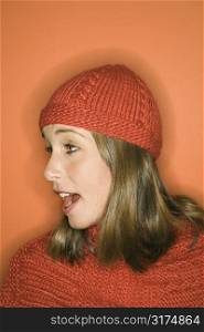 Side view of young adult Caucasian woman on orange background wearing winter hat and scarf.