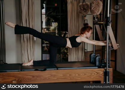 Side view of young athletic woman stretching legs on pilates machine or Cadillac reformer, wearing black top and trousers while exercising in gym or fitness studio, preparing muscles for training. Young ginger athletic woman exercising on pilates reformer in studio