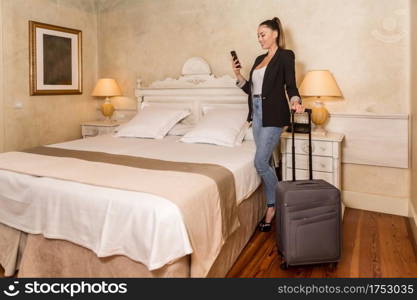 Side view of young attractive female standing with suitcase and using mobile phone in cozy bedroom. Business woman with smartphone in hotel room