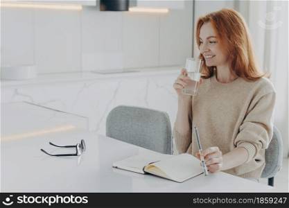 Side view of young beautiful red-haired smiling woman sitting in modern kitchen at home, drinking fresh water from glass and smiling while planning her day in morning, starting day with healthy habits. Young beautiful red-haired smiling woman sitting in kitchen at home, drinking fresh water from glass