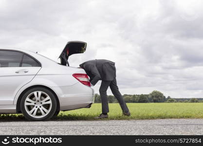 Side view of young businessman looking in trunk of car at countryside