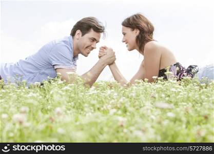 Side view of young couple arm wrestling while lying on grass against sky