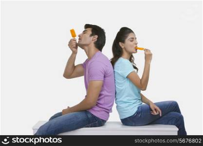 Side view of young couple enjoying ice lollies on bench over white background