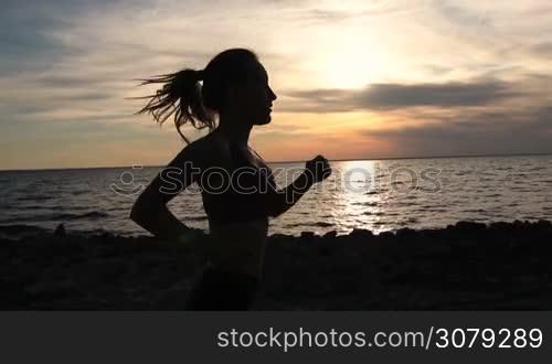 Side view of young fit female with ponytail running and exercising in the evening on the seaside. Silhouette of sporty runner woman jogging on the beach in twilight time. Slow motion. Steadicam stabilized shot.