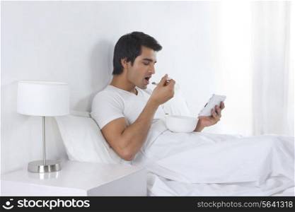 Side view of young man having breakfast while using digital tablet in bed