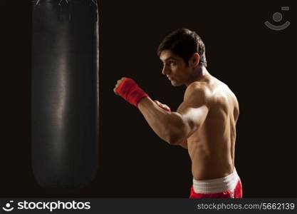 Side view of young man hitting punching bag isolated over black background