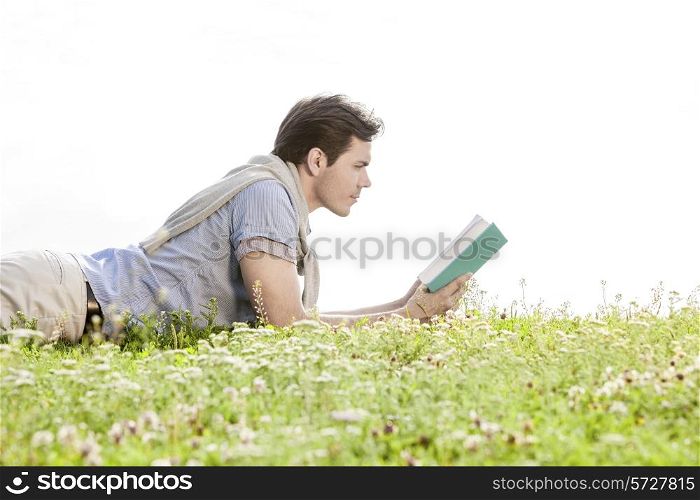 Side view of young man reading book while lying on grass against clear sky