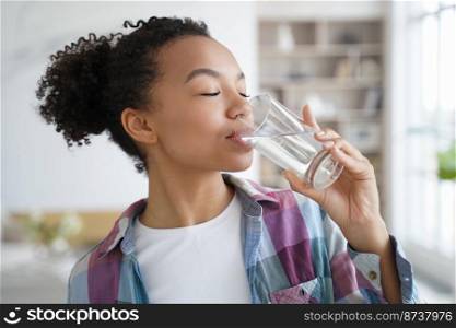 Side view of young mixed race girl drinking clean mineral still water from glass at home. Beautiful thirsty teen lady enjoying daily healthy lifestyle habit. Natural beauty, body skincare, wellness.. Young mixed race girl drinking clean mineral water at home. Healthy lifestyle habit, natural beauty