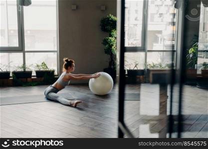 Side view of young red haired woman with perfect slim body sitting in split position on yoga mat in fitness studio bending forward and touching large stability exercise ball next to panoramic window. Sportive woman stretching legs in split position, training with large exercise ball