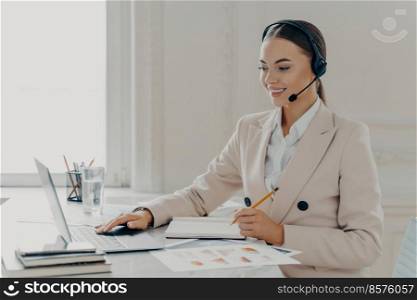 Side view of young smiling business woman wearing taking part in web conference, looking at laptop screen, call center operator consulting customer while sitting behind work desk in modern office. Call center operator wearing headset talking with client