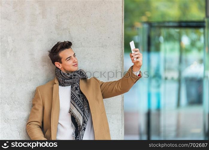 Side view of young trendy man leaning on a wall outdoors wearing denim clothes while taking a selfie in sunny day