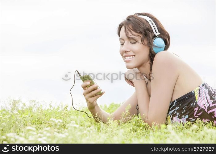 Side view of young woman listening to music through cell phone using headphones while lying on grass against clear sky