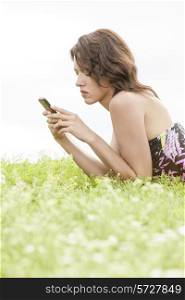Side view of young woman text messaging through cell phone while lying on grass against clear sky