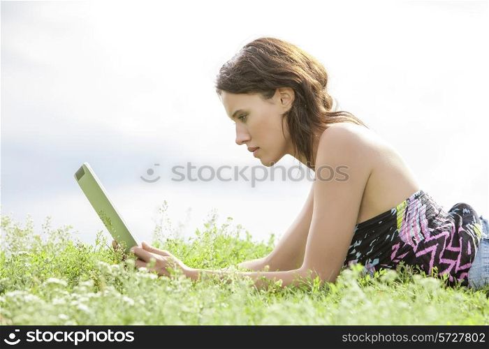 Side view of young woman using Tablet PC while lying on grass against sky