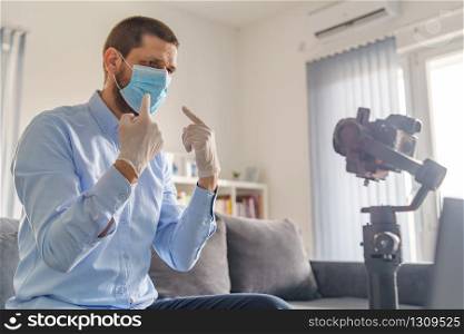 Side view on caucasian adult man blogger sitting at home in day with gimbal stabilizer camera making video about pandemic virus disease spread wearing protective mask gloves for prevention quarantine