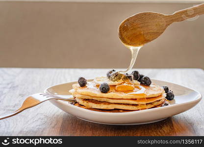 Side view on wooden spoon over the plate with pancakes and blueberries with butter pouring honey or maple syrup on the table