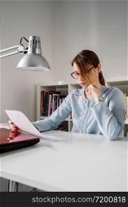 Side view on young adult caucasian woman female girl sitting by table at office work holding paper document reading analyze study got fired or bad report during the crisis economic recession in day