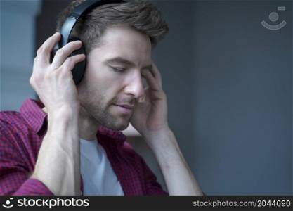 Side view photo of young pleased german man with closed eyes listening music in headphones with delighted face expression, austrian guy enjoying favourite playlist while spending free time at home. Young pleased german man with closed eyes listening music in headphones at home with delighted face