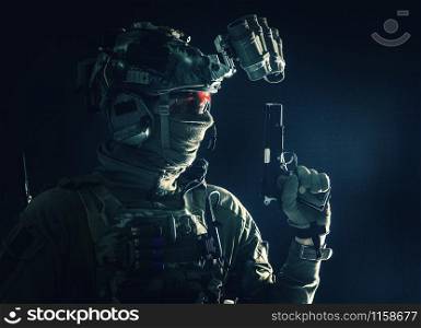 Side view portrait of army soldier, modern combatant, special forces fighter in helmet, night-vision device, radio headset, hiding identity behind mask, armed service pistol, low key studio shoot. Combatant armed with service pistol in darkness